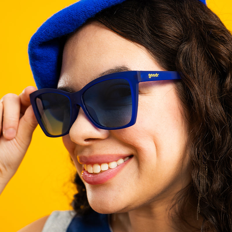A woman in blue angled cat-eye sunglasses with blue lenses and a blue beret smiles to the side.