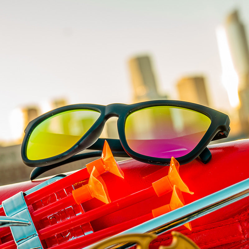 Three-quarter angle view of black sunglasses with reflective pink lenses sitting atop a dart gun, a city skyline behind it.