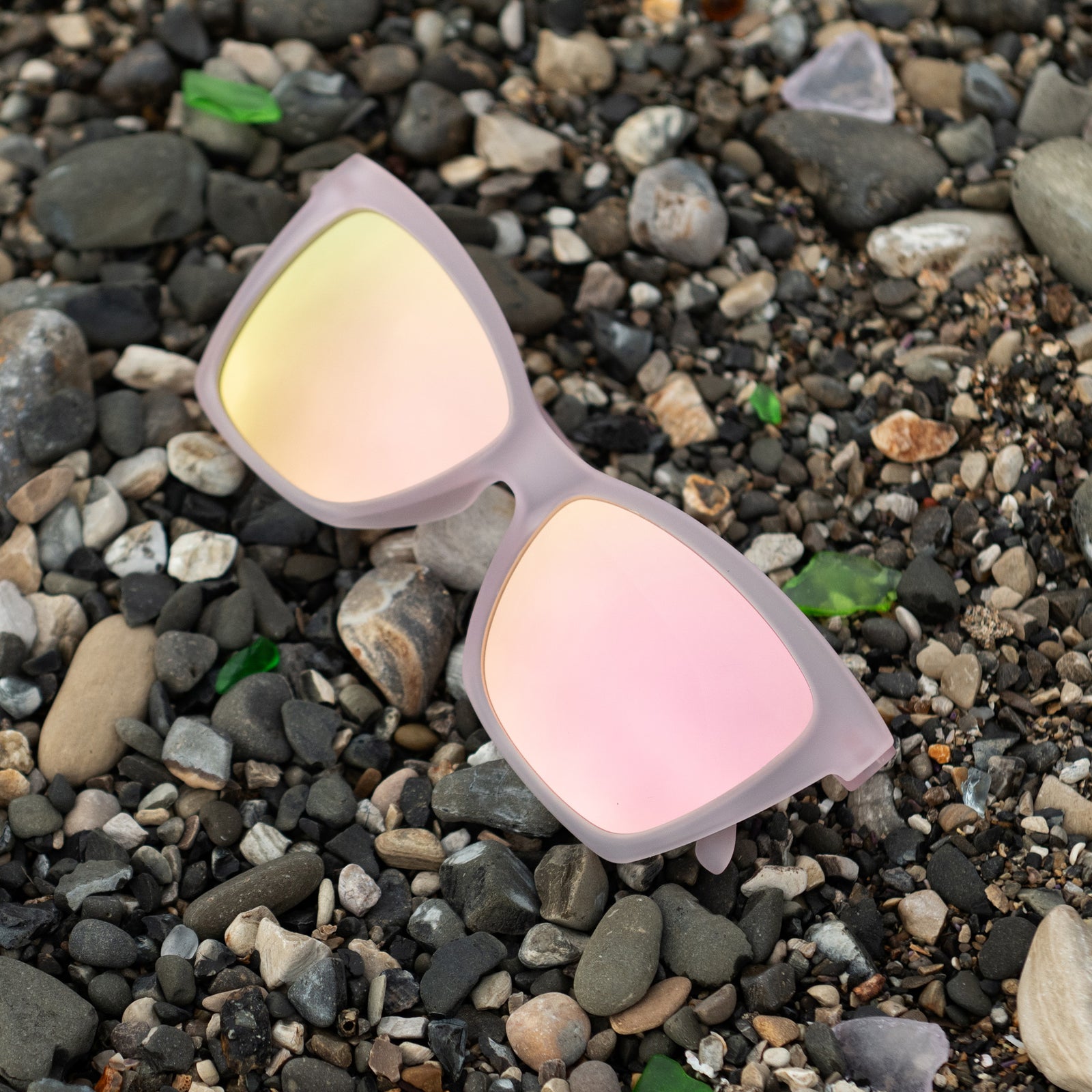 Translucent pink angular cat-eye Pop Gs with light pink reflective lenses lying on beach pebbles with sea glass.