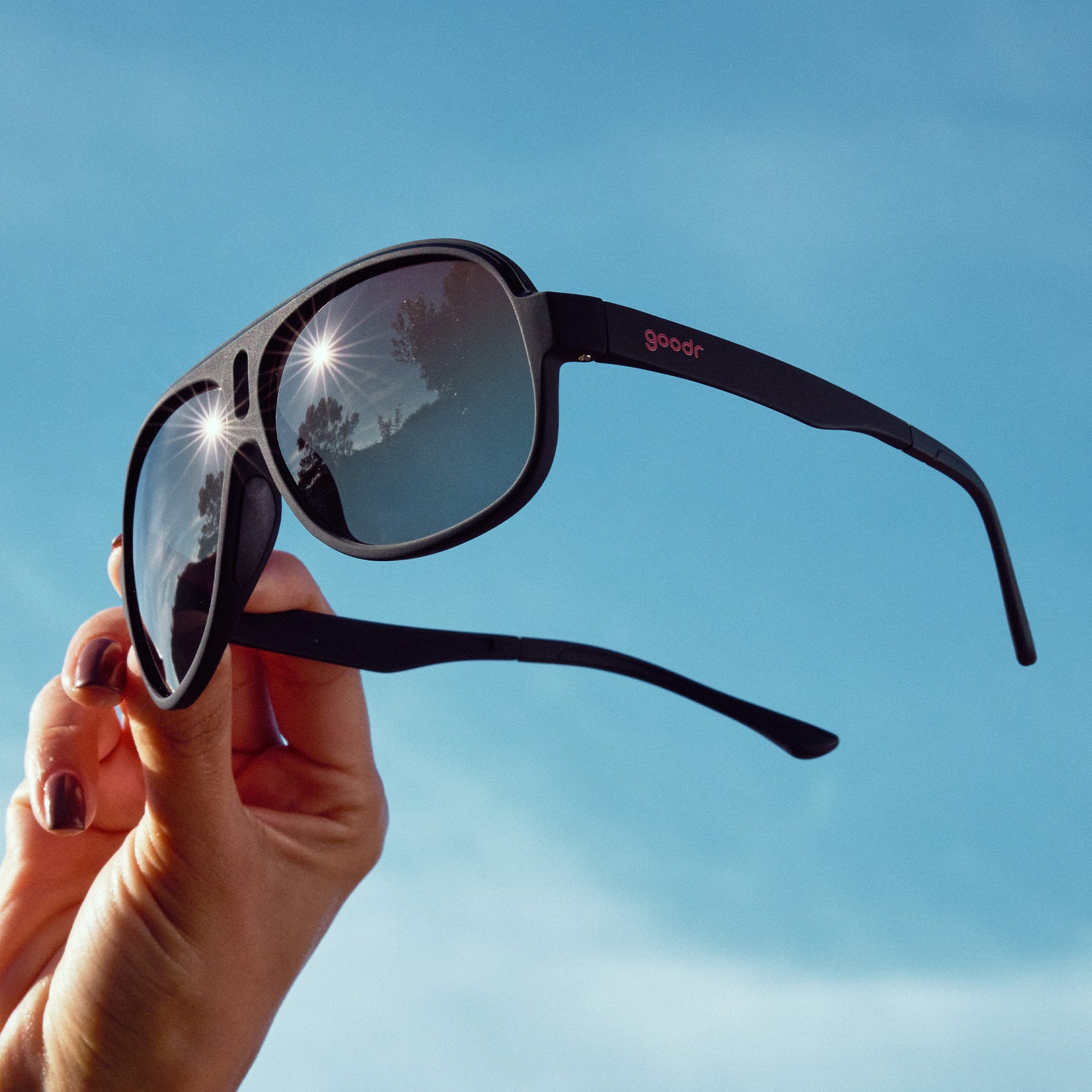 A manicured hand holds a pair of black aviator sunglasses with amber gradient lenses against the sky at a three-quarter angle.
