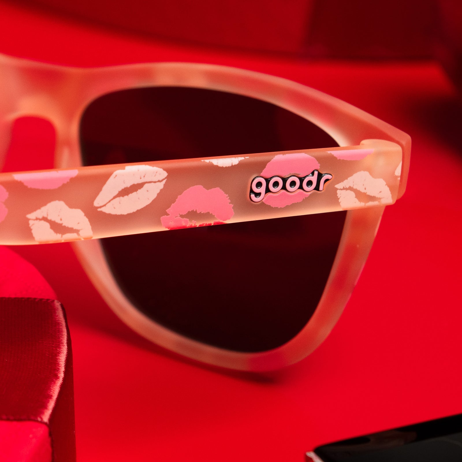 A pair of pale pink sunnies with a flirty all-over-kiss print is reflected in a vanity mirror that has been kissed with pink and red lipstick.