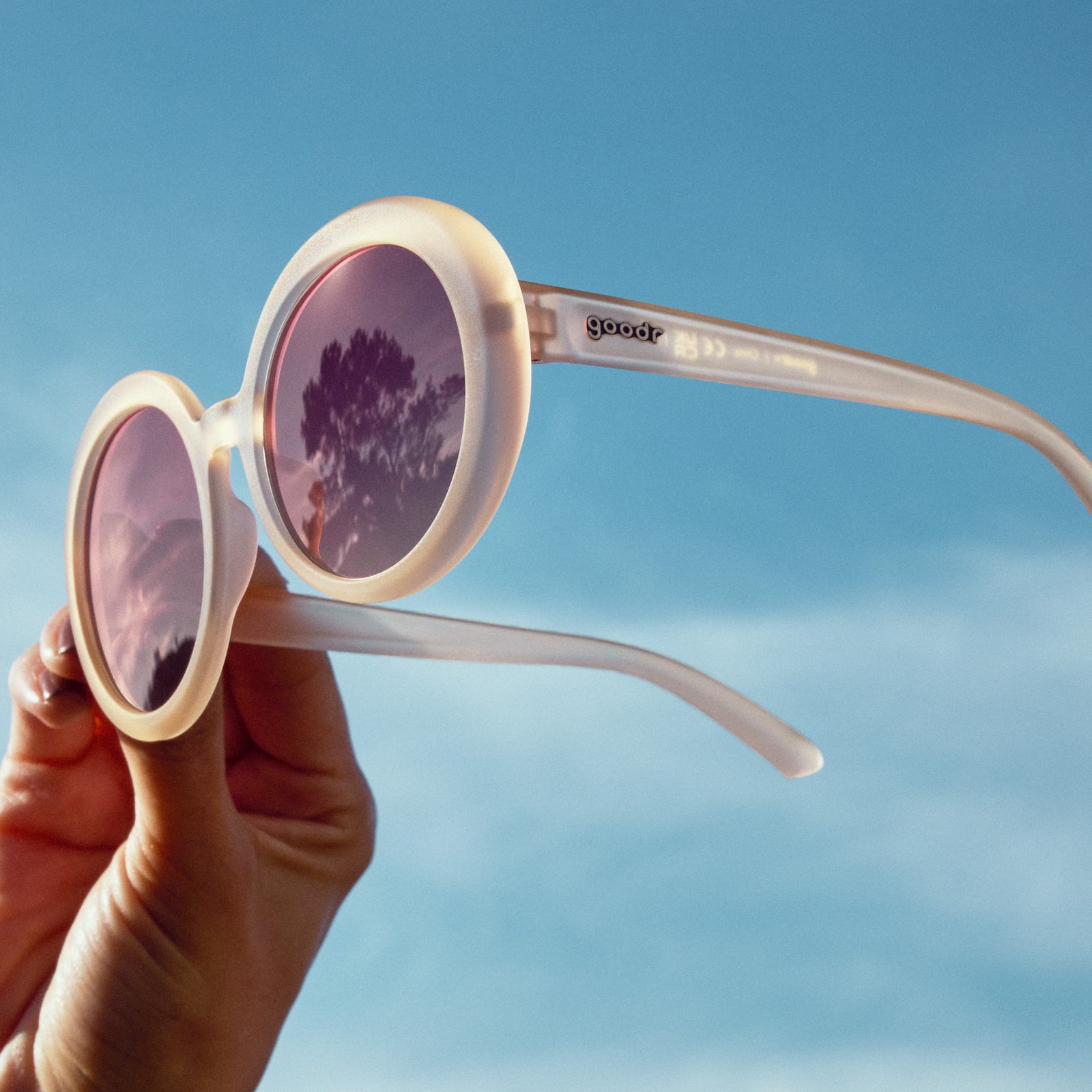 A hand holds up oversized, round beige sunglasses with purple gradient lenses against the sky.