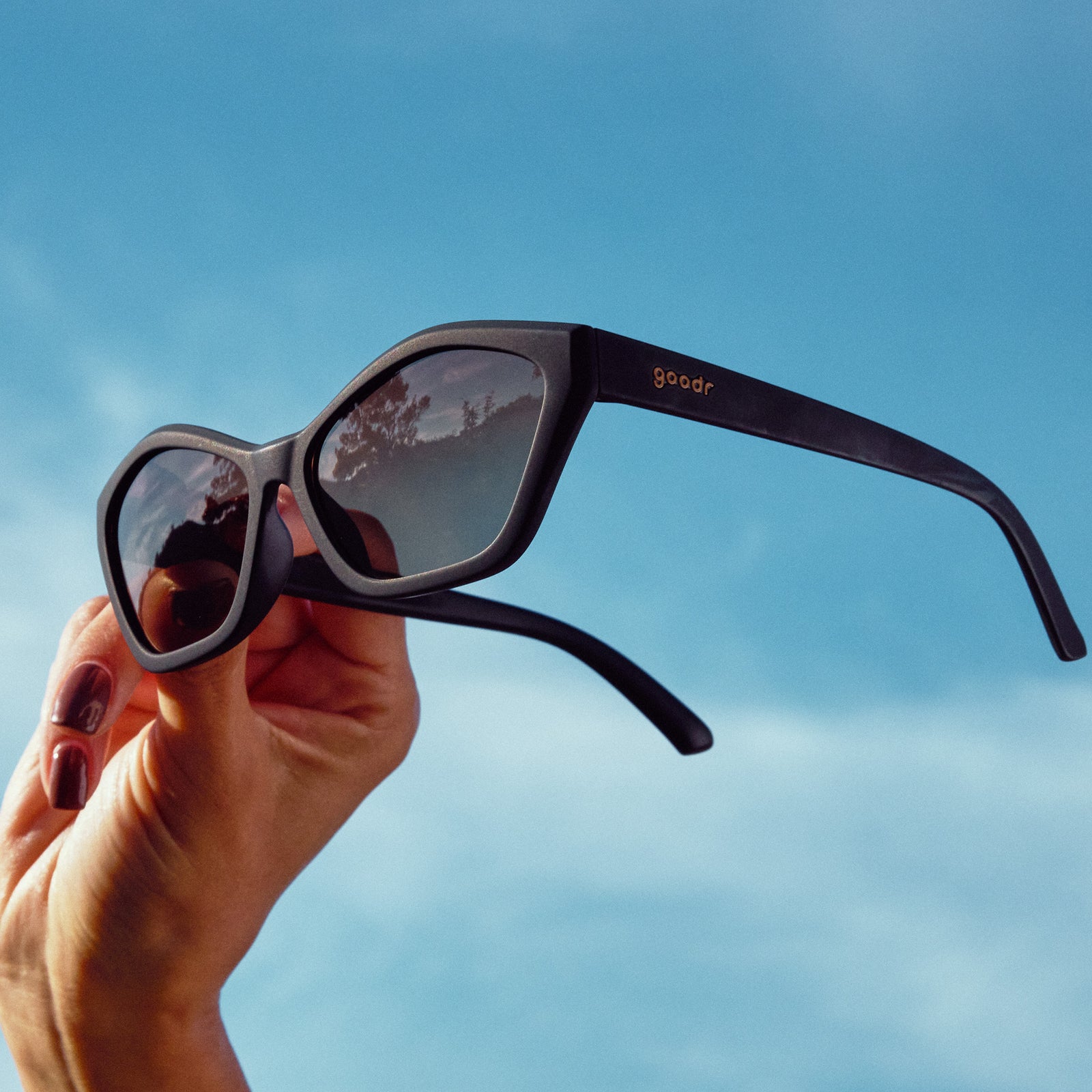 A hand holds up black geometric cat-eye sunglasses with amber gradient lenses against the sky at a three-quarter angle.
