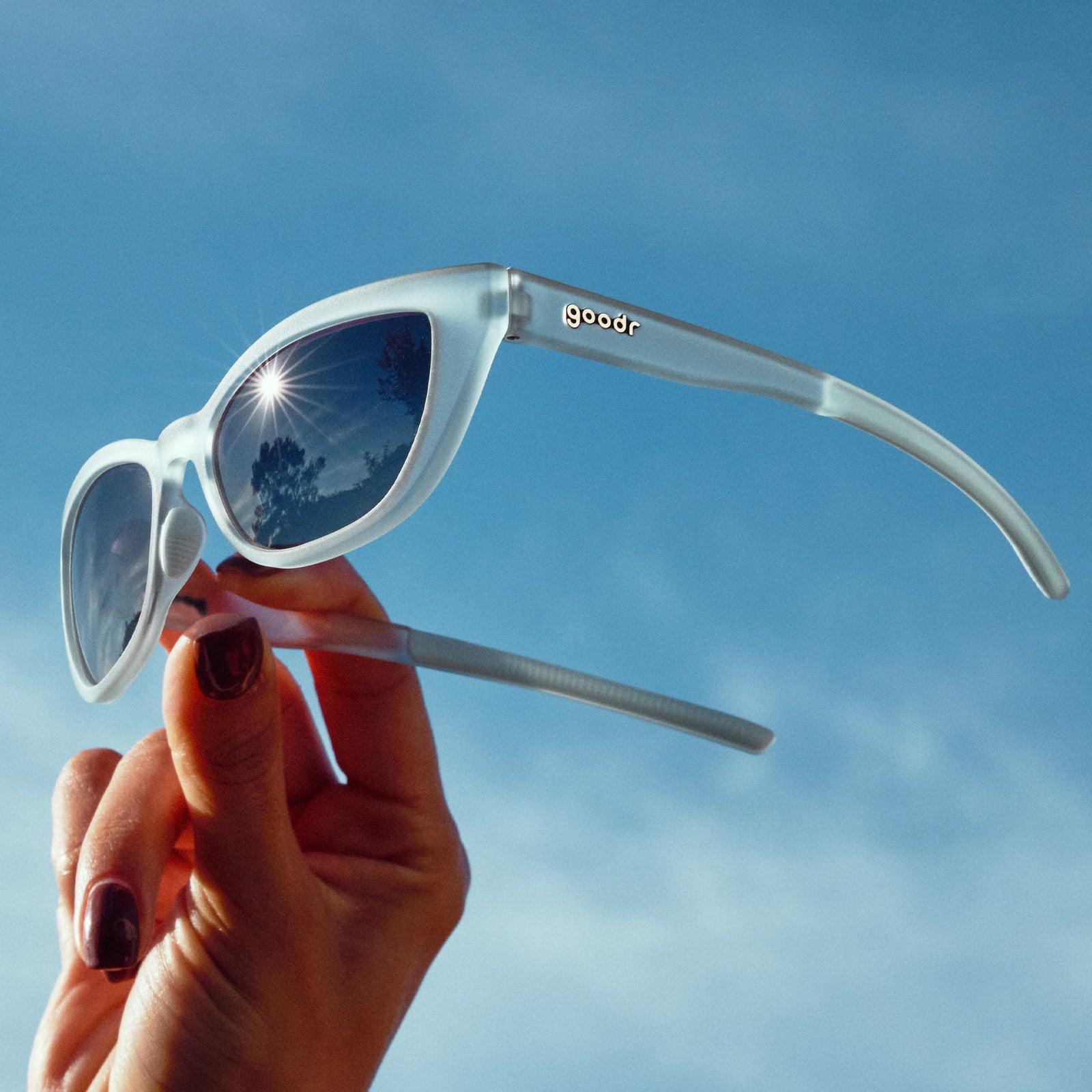 A manicured hand holds light blue cat-eye sunglasses with gradient violet lenses against the sky at a three-quarter angle.