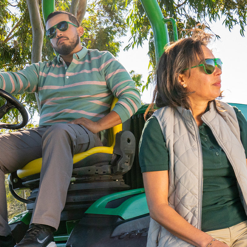 A man and woman wearing green aviator sunglasses with non-reflective lenses sit on a tractor and strike a pose.
