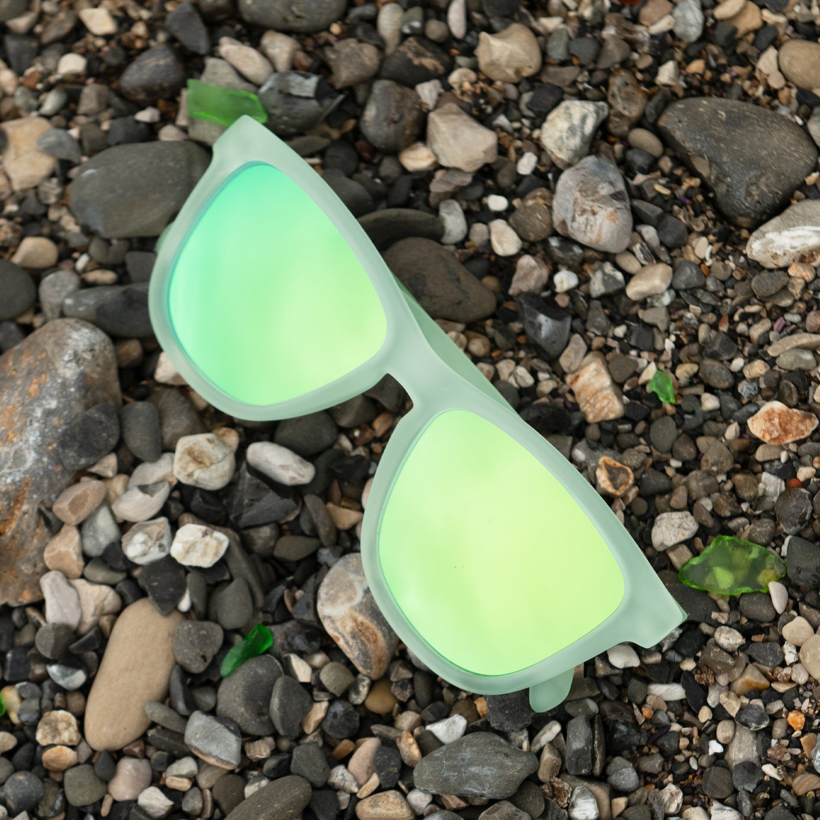 Translucent green OG sunglasses with green-blue reflective lenses lying atop beach pebbles with sea glass.
