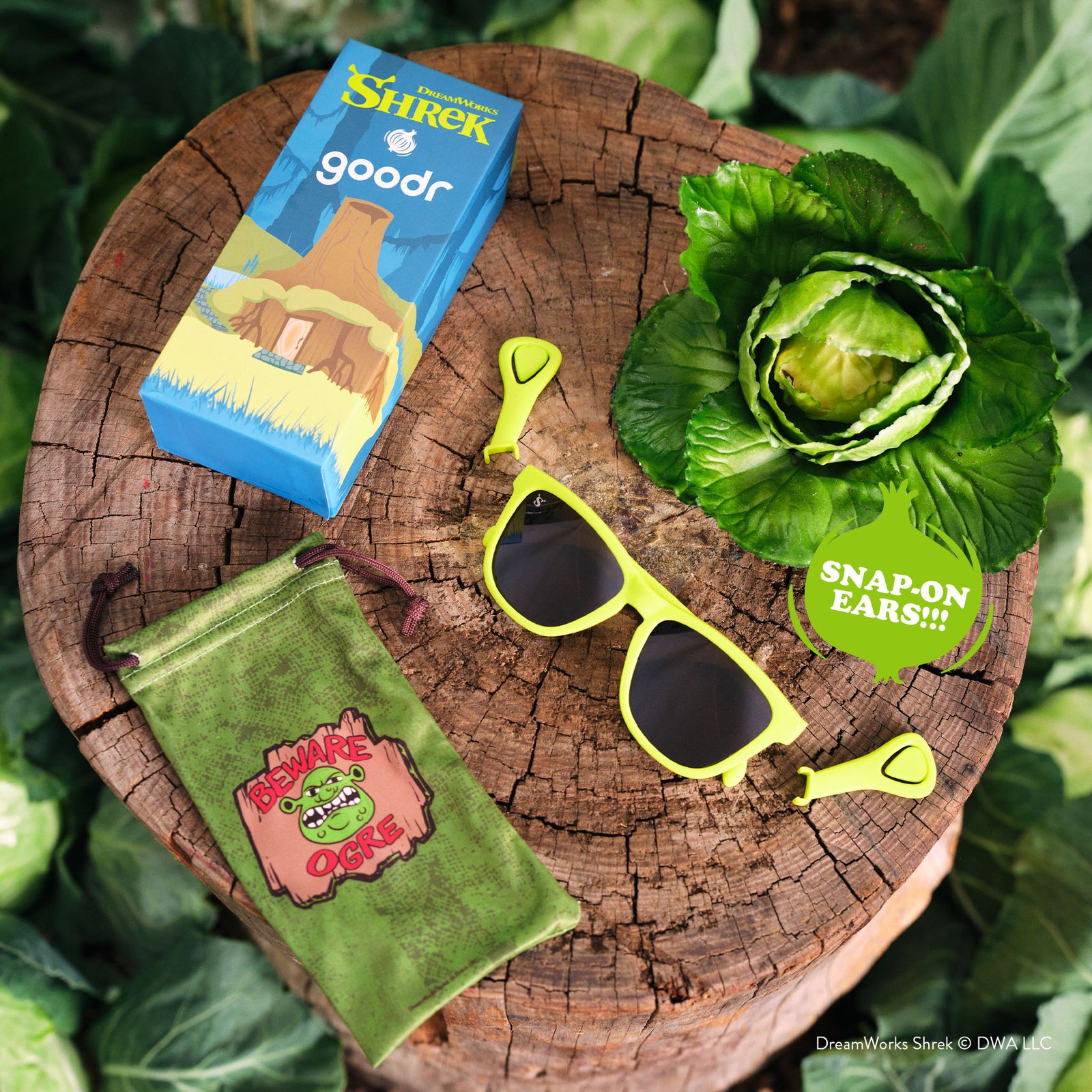 Goodr Shrek-themed sunglasses and packaging on a wooden stump with greenery.