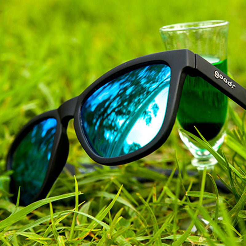 Three-quarter angle view of black sunglasses with reflective green lenses in a field, a glass of Absinthe behind them.