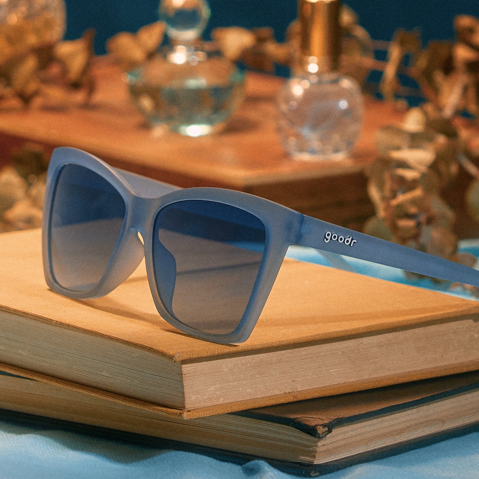 A pair of translucent, pale blue Pop Gs on top of some vintage books, dried flowers and perfume bottles in the background.