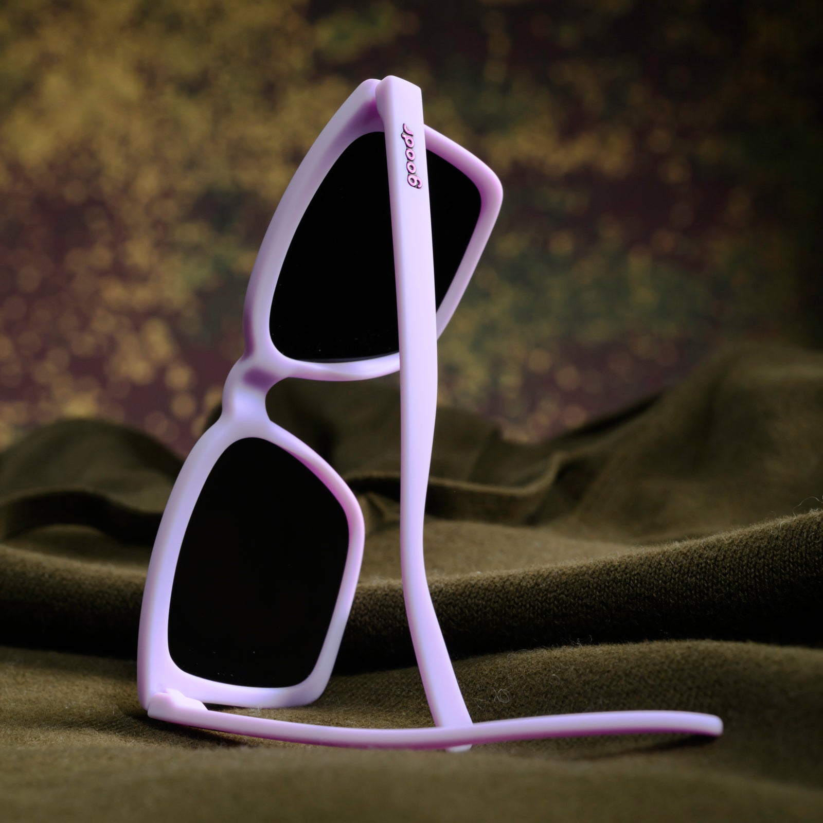 Close-up of a woman’s hand holding a pair of lavender square Slate G sunglasses against her black leather jacket.