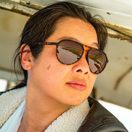 A woman in a bomber jacket and brown tortoiseshell aviator sunglasses looks out, an airplane hanger reflected in the lenses.