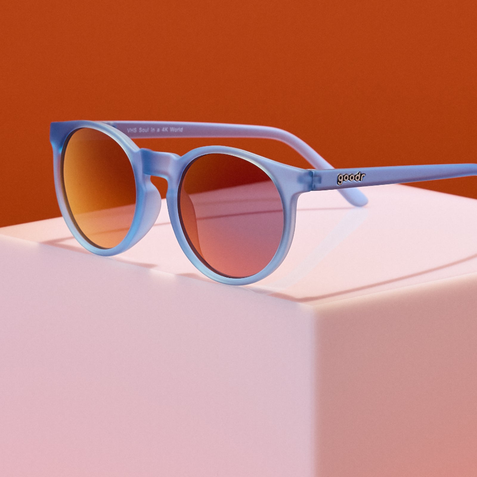 Blue sunglasses with red-tinted lenses on a pink surface against an orange background