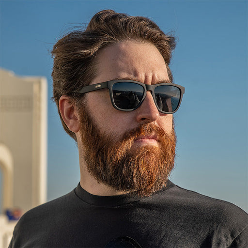 A redheaded white man wearing black sunglasses with black lenses sternly stares into the distance straight-faced.