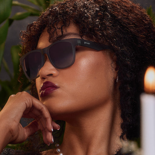 Close-up of a young woman with plum-lacquered lips in dark gray sunglasses, her hand lifted thoughtfully to her chin.