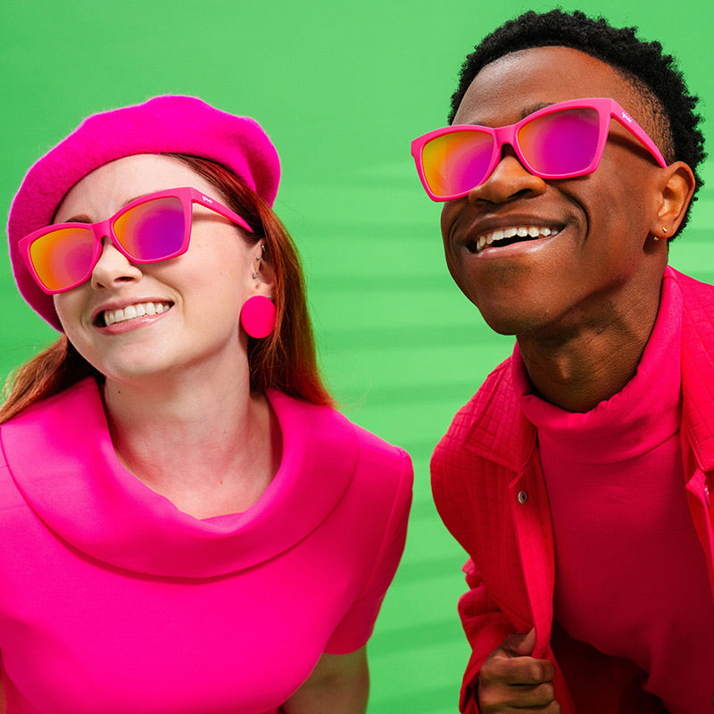 A man and woman in vibrant pink outfits wear bright pink angled cat-eye sunglasses with pink reflective lenses.