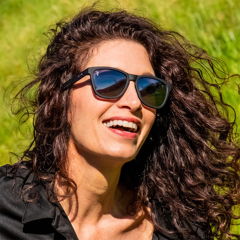 A brunette woman wearing black sunglasses with black lenses smiles on a sunny day, a golf course behind her.