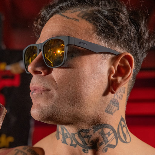 A tattooed strong man wearing large square-shaped black sunglasses with mirrored amber lenses looks off the side.