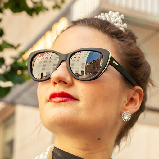 A royal woman with large diamond earrings wearing black cat-eye frames with gold trim and black gradient lenses.