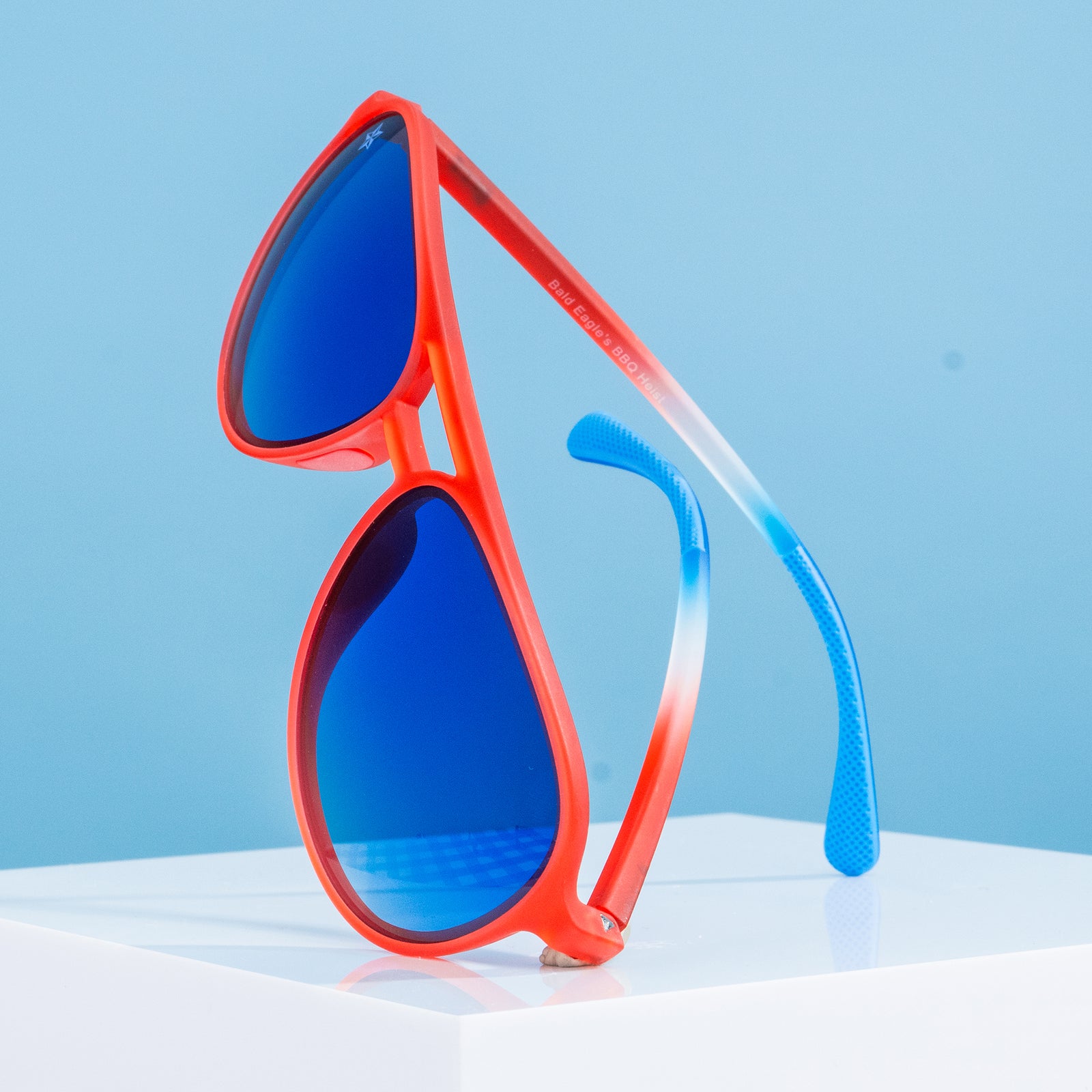 Three-quarter angle view of red, white,+blue aviator sunglasses set upright. Blue gradient lenses. Right lens star etch.