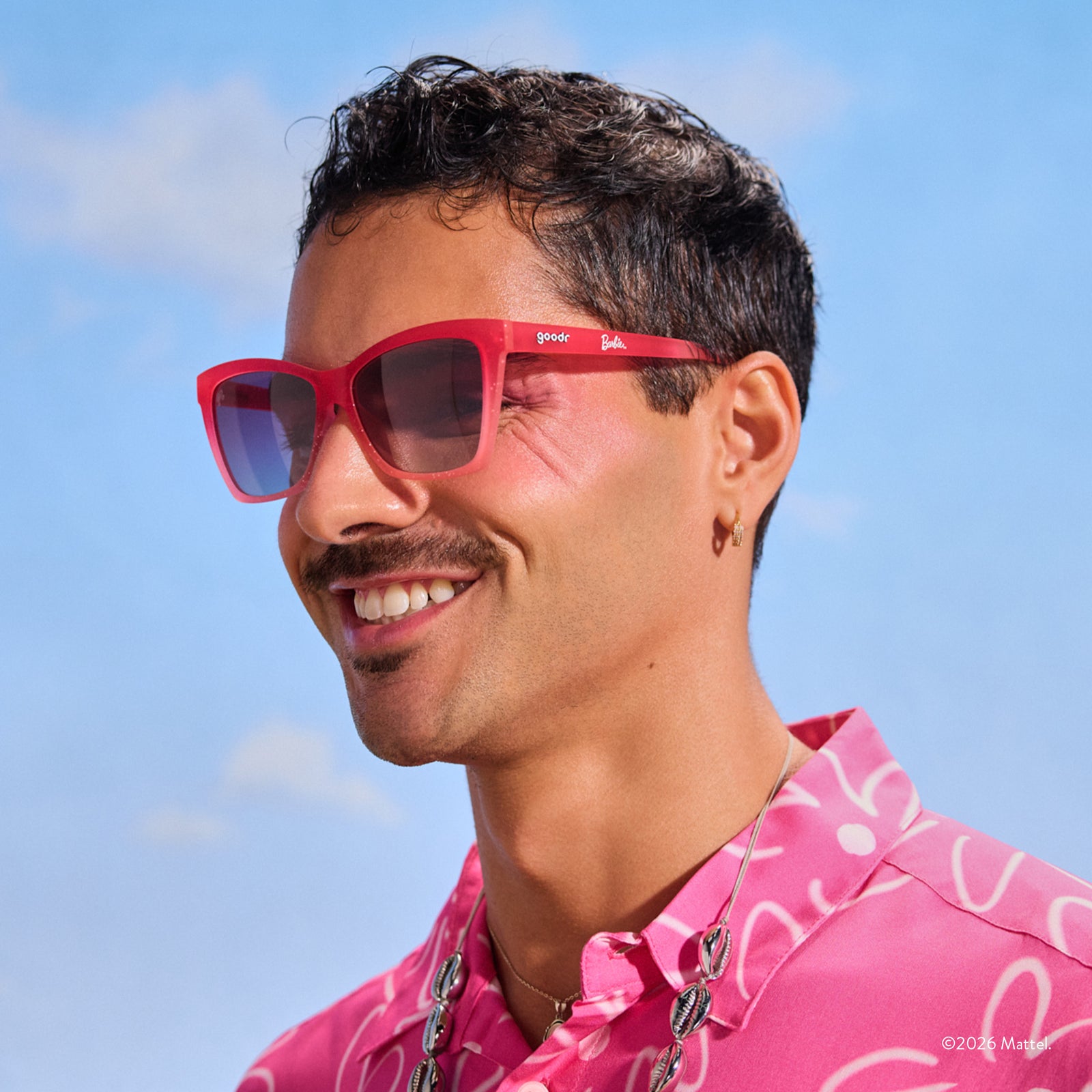 A young man with dark hair wears a pair of translucent, glittery pink ombre Pop G sunglasses against a sky-like background.