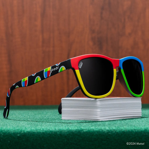 Three-quarter view of a pair of UNO shades perched atop a deck of UNO cards, on a green game table in front of wood paneling.