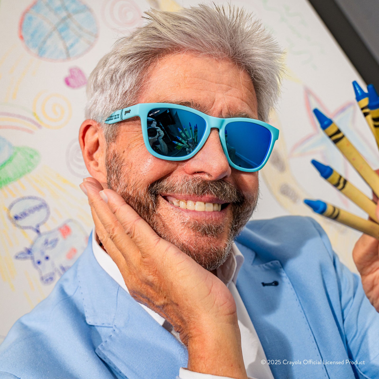 A smiling gray-haired man wears Blizzard Blue Crayola shades & holds crayons in front of a white sheet with drawings