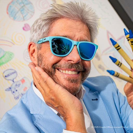 A smiling gray-haired man wears Blizzard Blue Crayola shades & holds crayons in front of a white sheet with drawings