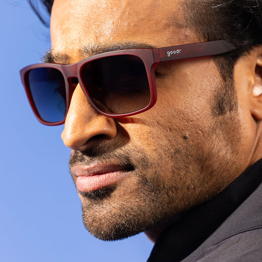 A stylish man with dark hair smirks, wearing dark red Grand G frames and black gradient lenses.