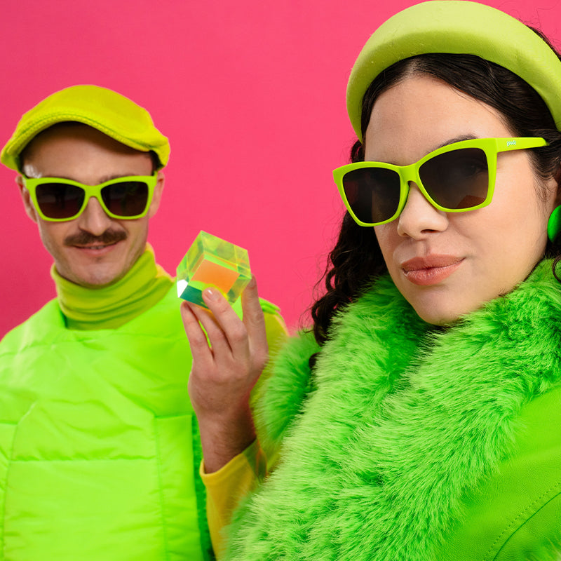 A man and woman in neon green outfits wear lime green angled cat-eye sunglasses with black gradient lenses.