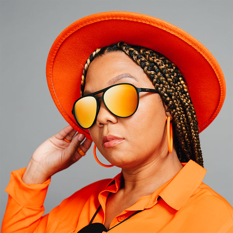 A woman poses fiercely in an orange hat and shirt wearing black aviator sunglasses with amber lenses.