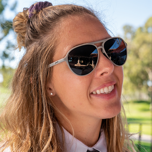 A smiling woman in a collared shirt at a golf clubhouse wears gray aviator sunglasses with non-reflective lenses.