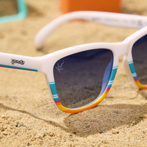 A pair of white shades with a striped, beach-inspired design and blue lenses sits in the sand. Lenses have a Ken™ lens etch.