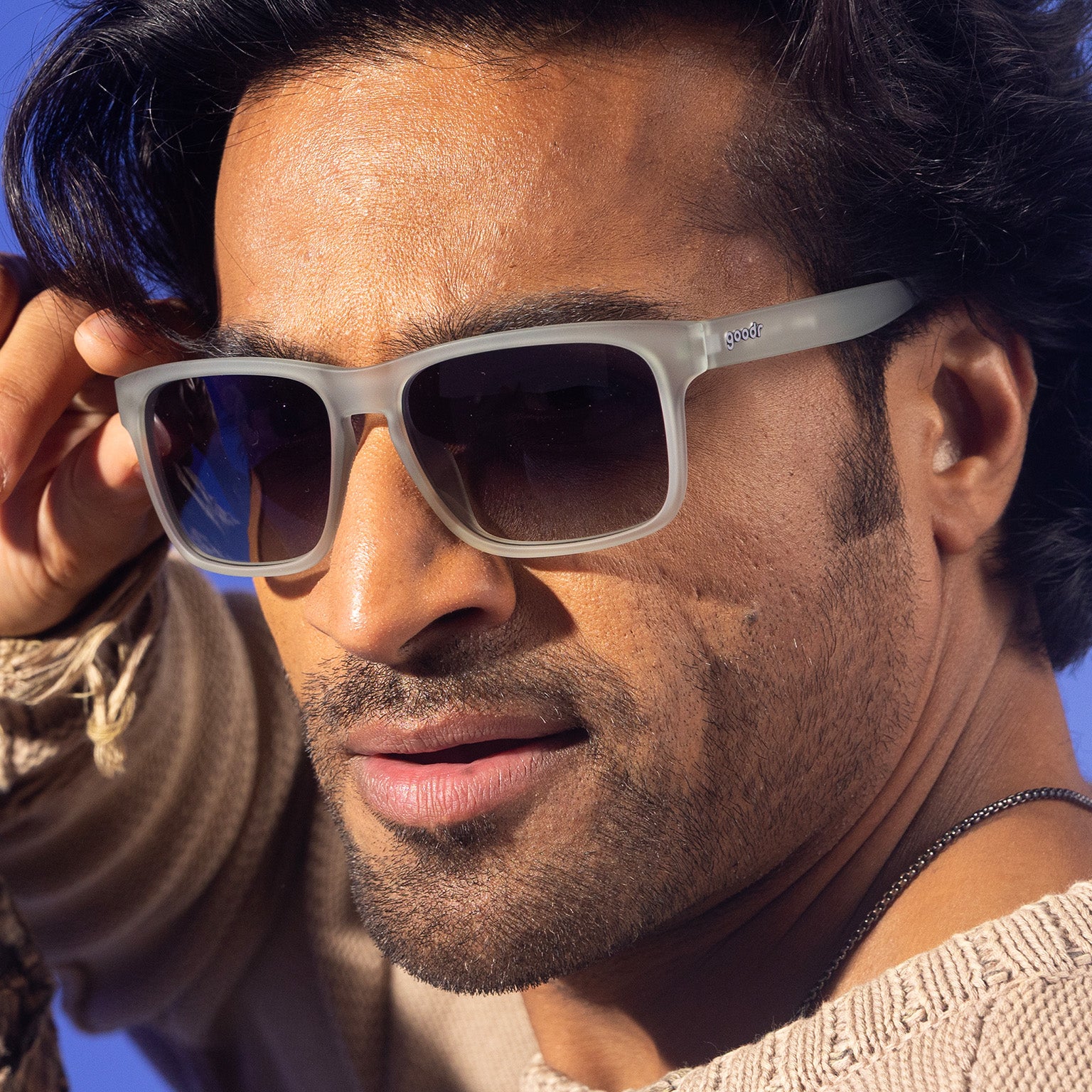 A sleek, stylish man with wavy black hair playfully holds the temple of his translucent gray Grand Gs with black lenses.