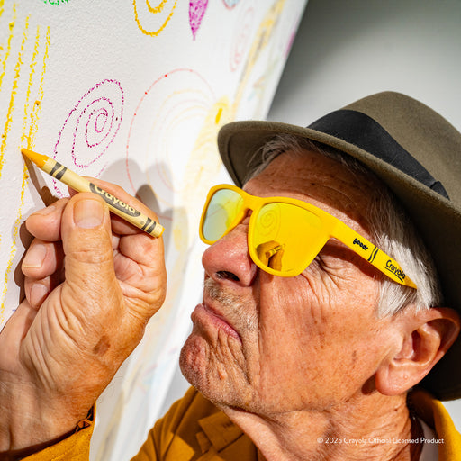 An older man in a fedora wearing Crayola Dandelion sunglasses concentrates while drawing something on a white wall
