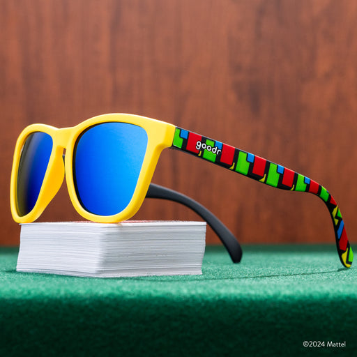 Three-quarter view of a pair of UNO shades perched atop a deck of UNO cards, on a green game table in front of wood paneling.