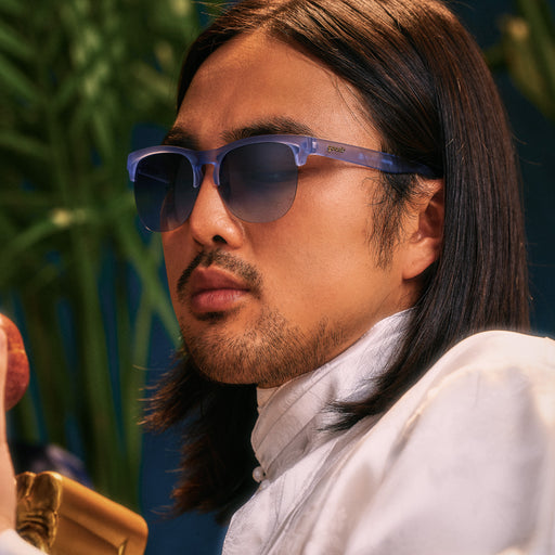 A young man with dark, shoulder-length hair poses in a pair of dusty blue, translucent Avant Gs, with palm fronds in the background.