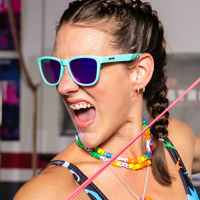 A woman with rainbow plastic necklaces and baby blue sunglasses with purple reflective lenses grunts ferociously at a gym.