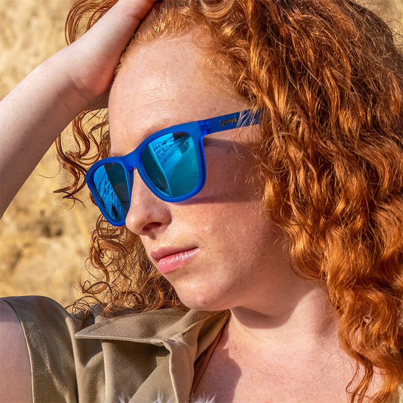 A woman wearing bright blue sunglasses with reflective blue lenses leans her head onto her hand, looking into the distance. 