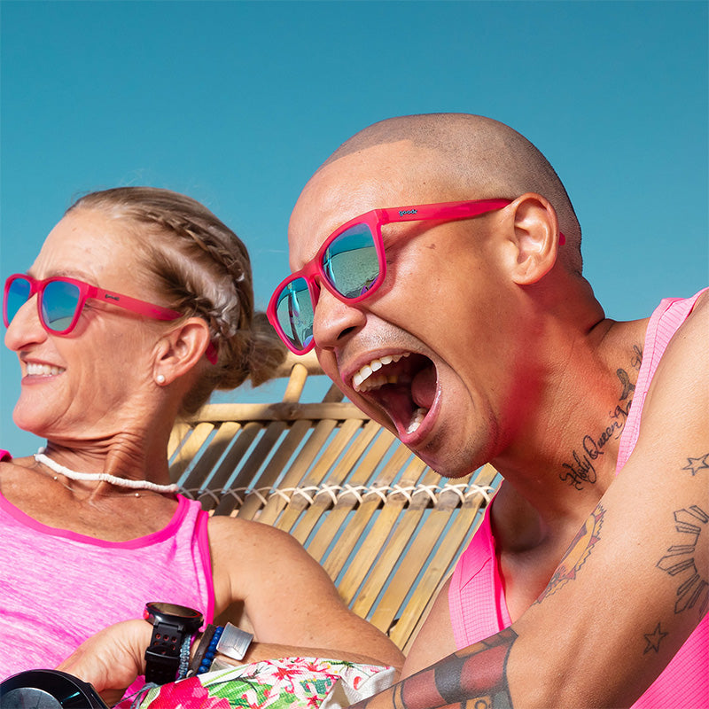 A man and woman wearing hot pink sunglasses with green lenses laugh while lounging on a tropical beach.