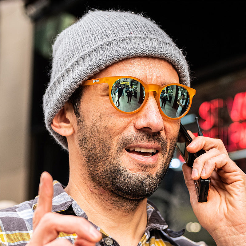 A hipster man in a gray beanie and polarized round orange sunglasses looks off to the distance while talking on a flip phone.