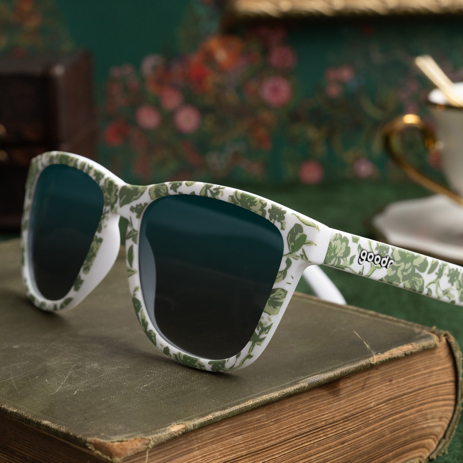 Three-quarter angle view of sunglasses with a green and white floral toile and green gradient lenses on top of an old book.