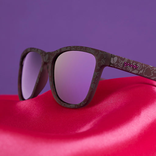 Three-quarter angle view of limited-edition sunglasses with black "self-love" patterned frames and purple polarized lenses on a pink satin bedsheet.