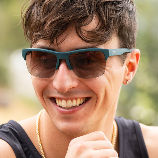 Close-up of a man outdoors, laughing with his hand near his mouth while wearing solid green half-rim sunglasses with polarized rose gradient lenses.