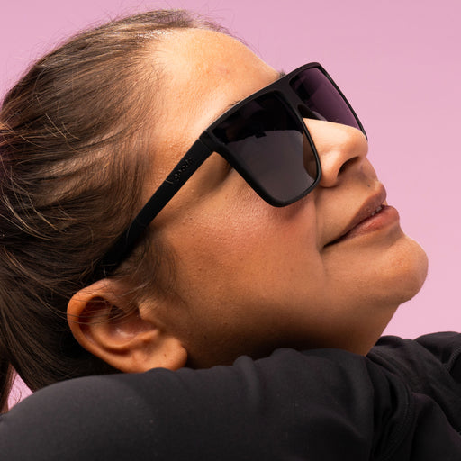 A closeup of a woman smiling, looking up in oversized black flat-top glasses with black non-reflective lenses.