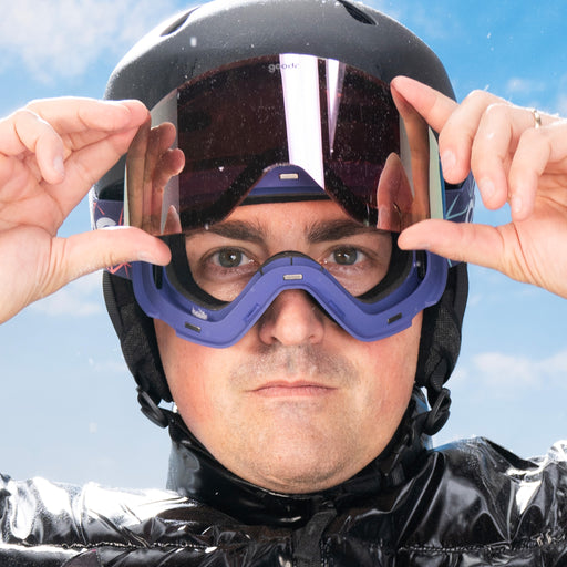 A man in black snow gear is showcasing the magnetic lens securing system by popping the reflective lens off the snow goggles.