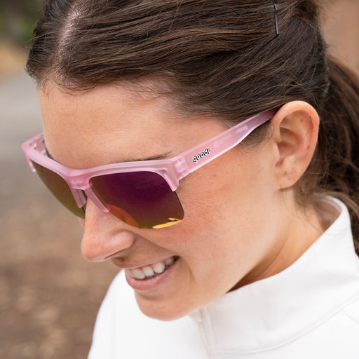 High angle of a woman’s side profile as she smiles while wearing half-rim translucent pink sunglasses with pink reflective polarized lenses.