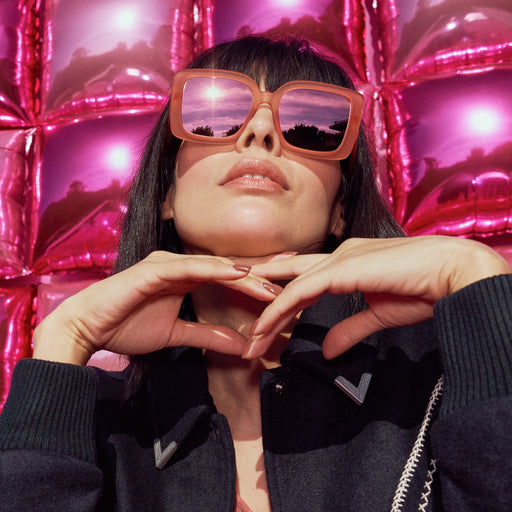 Portrait of a woman wearing oversized square sunglasses in coral pink with champagne gold reflective lenses in front of a shiny pink balloon wall.