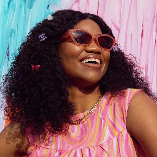 A smiling woman wearing geometric sunglasses with translucent coral pink frames and gradient lenses in front of a pastel backdrop.