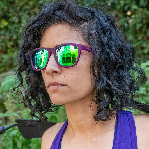 A woman in a garden fiercely stares off wearing purple sunglasses with green reflective lenses, holding a purple garden trow.