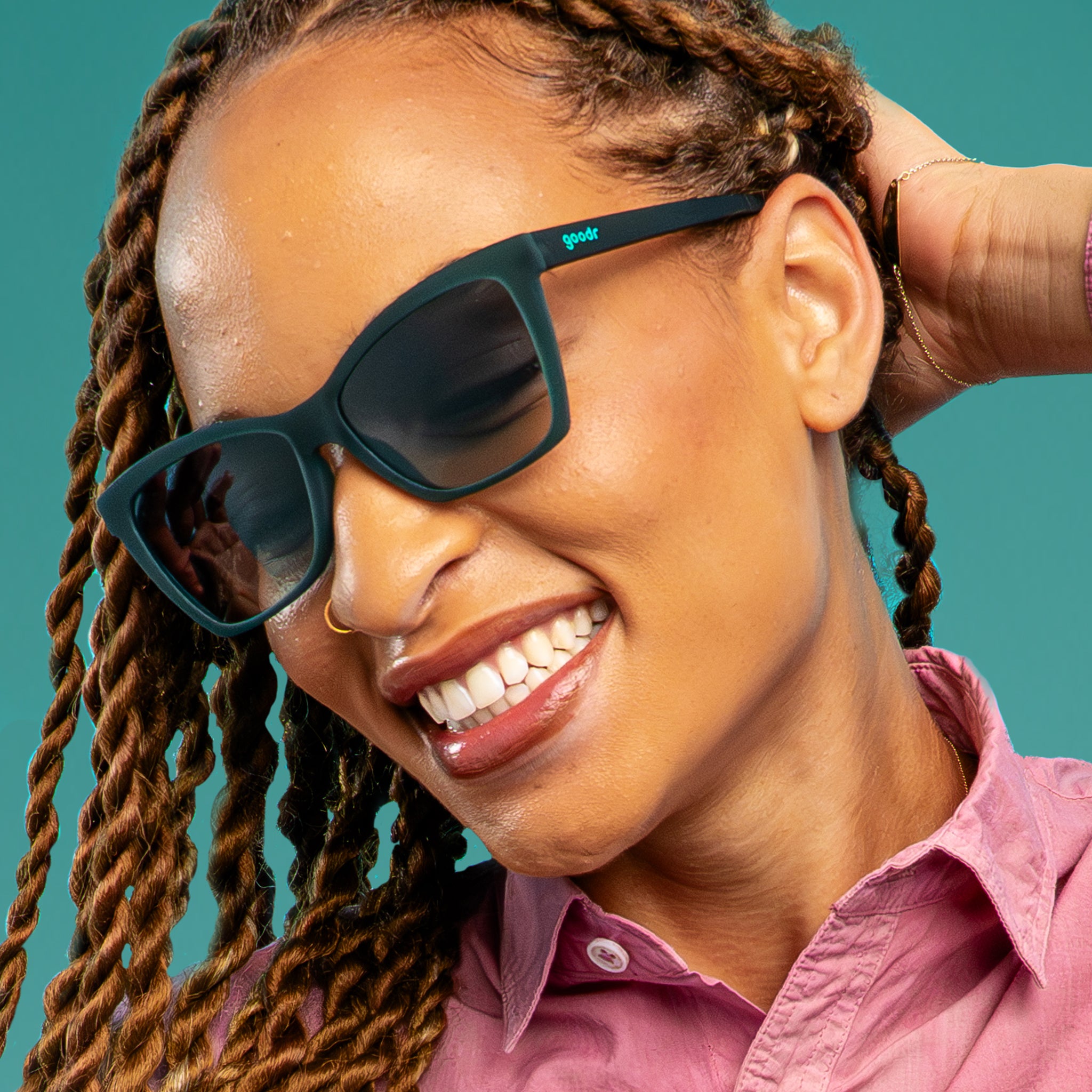 A woman with copper twist braids smiles confidently in a pair of translucent teal Pop Gs with green gradient lenses, posing against a teal backdrop.